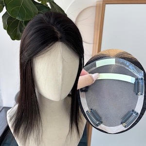 以下が含まれることがあります： マネキンの頭に展示された、自然な分け目の黒いヘアートッパー。画像には、固定用のクリップと粘着ストリップが示されたトッパーの裏側も表示されています。髪はストレートで、肩まで届いています。
