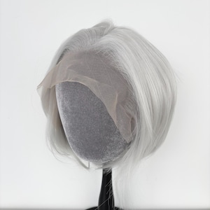 Puede incluir: Peluca corta gris plateado con frente de encaje, exhibida sobre una cabeza de maniquí gris. La peluca tiene un corte romo y cabello liso y fino. El frente de encaje es de color beige claro. Montada sobre un soporte negro sobre fondo blanco.