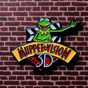 May include: A colorful pin featuring Kermit the Frog holding a camera, with the words "MuppetVision 3D" in red and white on a yellow banner. The pin is set against a brick wall background.
