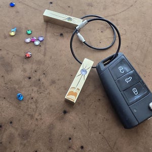 Puede incluir: Un llavero dorado con un cordón negro y dos abalorios rectangulares. Cada abalorio tiene una piedra transparente en el centro con un diseño de estrella. El llavero está unido a una llave de coche negra.