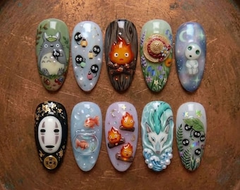 May include: A set of ten almond-shaped press-on nails featuring detailed 3D character art and illustrations inspired by animated films. Designs include a grey forest creature, soot sprites, a fire spirit, a straw hat, a white forest spirit, a white mask, goldfish in a bowl, and a white dragon with blue waves.