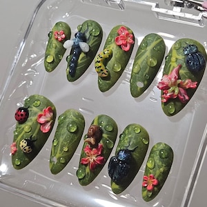 May include: A set of ten almond-shaped artificial nails. Each nail features a green leaf design with water droplets and miniature insect and floral embellishments. The insects include a dragonfly, ladybug, and beetle. The flowers are pink.