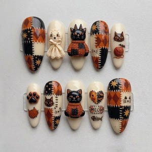 May include: A set of ten press-on nails featuring a cream base with orange and black patchwork patterns, 3D cat figures in sweaters, a small doll-like figure with a bow, paw prints, yarn balls, and the word MEOW written in black lettering.