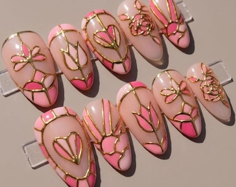May include: A set of ten almond-shaped press-on nails featuring a light pink base with vibrant pink accents and raised gold metallic lines forming intricate floral, heart, and geometric patterns across the surface of each individual nail.