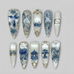 May include: A set of ten long, almond-shaped artificial nails. Each nail features a white base with intricate blue and gold designs, including floral patterns, a hummingbird, and a boat scene. The nails are arranged on a light gray surface.