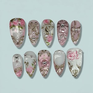May include: A set of ten artificial nails with a light pink and clear base. Each nail features intricate designs, including gold accents, bows, roses, and decorative embellishments. The nails are arranged on a light blue surface.