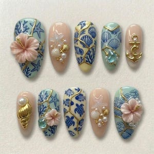 May include: A collection of ten artificial nails with a seaside theme. The nails are decorated with seashells, starfish, and hibiscus flowers in blue, pink, and gold. Some nails have 3D decorations.