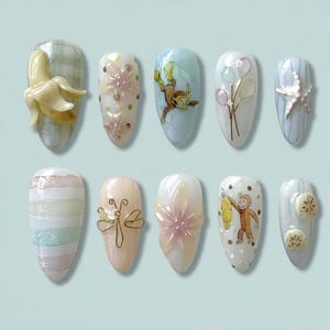 May include: Ten almond-shaped nail designs are displayed. The designs feature a 3D banana, floral accents, a monkey with a banana, balloons, a starfish, stripes, a dragonfly, and fruit slices. The nails are painted in pastel shades.