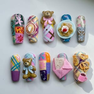 Pode incluir: Um conjunto de dez unhas postiças com vários designs 3D, incluindo ursinhos de peluche com laços, um prato de comida, um envelope cor-de-rosa, pretzels e uma caneca de café, além de padrões coloridos como xadrez, flores e patchwork.