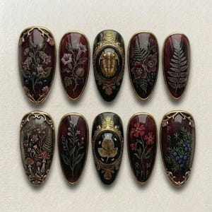 May include: A set of ten oval-shaped, dark burgundy nail art designs with intricate floral, fern, and insect motifs. Each nail features a gold-colored border. Some nails have a gold-colored embellishment in the center.