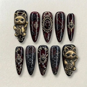 May include: A set of ten long, almond-shaped press-on nails. The nails feature a dark red base with black rose and lace designs, gold embellishments, and two nails with a gold cat charm. The nails are a gothic, elegant style.