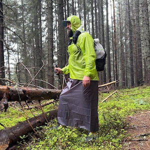 May include: A person wearing a neon yellow rain jacket with a hood, a gray skirt, and a backpack hikes through a forest. The person is using a hiking pole. The forest floor is covered in green plants.