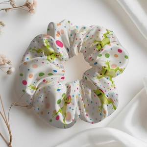 May include: White scrunchie with a playful frog and polka dot pattern. The scrunchie features green frogs and colorful dots on a white background. A stylish hair accessory for everyday wear.