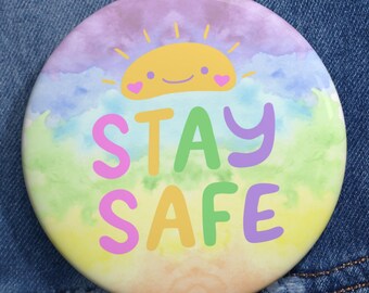 Può includere: Un bottone rotondo con un design colorato e la scritta "STAY SAFE". Il bottone presenta un sole sorridente e uno sfondo arcobaleno. Il bottone è appuntato su una giacca di jeans blu.
