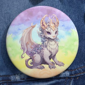 May include: A round button featuring a fantasy dragon illustration. The dragon is depicted in shades of purple, white, and gray, with a colorful watercolor background of blues, greens, and purples. The button is set against a denim jacket.