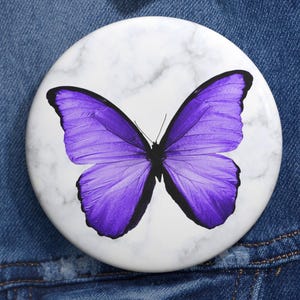 May include: A round white button with a marble-like pattern and a vibrant purple butterfly design. The butterfly has detailed wing patterns and is centered on the button. The button is set against a denim jacket.