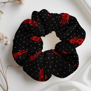 May include: Black scrunchie with a repeating pattern of red handprints and small white polka dots. The hair accessory is made of a soft fabric and is designed to hold hair in place. A stylish hair accessory.
