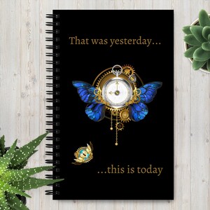Puede incluir: Cuaderno negro con espiral con el texto "That was yesterday... ...this is today." La portada presenta un diseño steampunk con un reloj, engranajes y alas de mariposa azules. También hay un pequeño emblema dorado y azul.