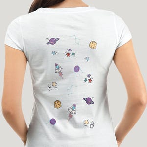 Op de afbeelding: Wit T-shirt met een kleurrijk ruimte-thema ontwerp. Het ontwerp bevat planeten, sterren, raketten en sterrenbeelden in paars, geel, rood en blauw. Het shirt heeft een ronde hals en korte mouwen.