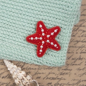 Puede incluir: Una aplicación roja en forma de estrella de mar con perlas blancas está adherida a una tela de punto verde claro. La estrella de mar está adornada con pequeñas cuentas rojas en los bordes. Se ve una concha en la parte inferior de la imagen.