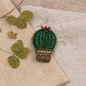 Op de afbeelding: Een groene kralen cactus broche met een rode bloem bovenop, in een bruine kralen pot. De broche is op een beige stoffen achtergrond.