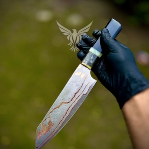 May include: A handcrafted chef's knife with a patterned blade and a dark handle. The blade has a unique design with reddish-brown streaks. The handle is a dark blue color with a light green accent. The knife is held by a gloved hand.