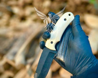 May include: A black blade knife with a cream-colored handle. The handle has three dark rivets. The knife is held by a gloved hand. The background is blurred, showing a pile of wood.