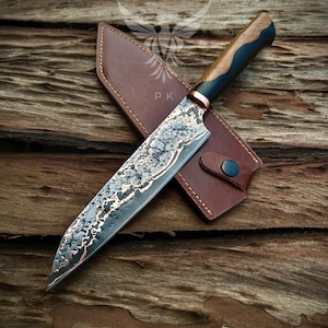 May include: A handcrafted chef's knife with a patterned blade and wooden handle, resting on a brown leather sheath. The blade has a unique, textured design with copper accents. The handle features a dark blue and wood grain design.