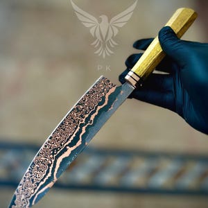 May include: A chef knife with a patterned Damascus steel blade featuring copper and dark grey layers. The handle is made of light-colored wood with a metallic bolster. A logo with a bird icon and the letters PK is visible above the blade.