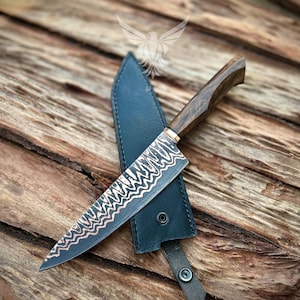 Könnte beinhalten: Ein handgefertigtes Kochmesser mit einer dunklen, gemusterten Klinge und einem Holzgriff. Das Messer liegt auf einer schwarzen Lederscheide mit Druckknopfverschluss. Die Klinge hat ein dekoratives geätztes Design mit Kupferakzenten.