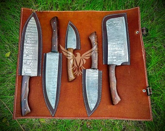 May include: A set of five Damascus steel knives with wooden handles and leather sheaths, displayed on a brown leather roll. The blades have a unique patterned design. The set includes a cleaver, a chef's knife, and smaller utility knives.