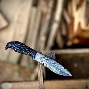 May include: A decorative knife with a dark handle and a patterned blade. The handle has a textured design, and the blade features an intricate etched pattern. The knife is displayed on a wooden stand, with a blurred background.