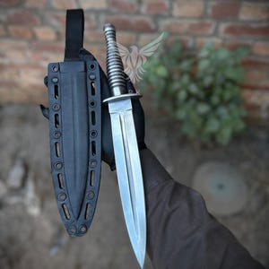 Puede incluir: Una daga plateada con un mango acanalado y una hoja de doble filo se muestra junto a su funda negra. La funda tiene una trabilla para el cinturón y varios ojales. El fondo es una pared de ladrillos y vegetación.