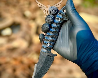 May include: A tactical knife with a curved, black blade and a handle featuring blue and black accents. The handle has a textured pattern. The knife is held by a gloved hand, with a logo of a bird in the background.