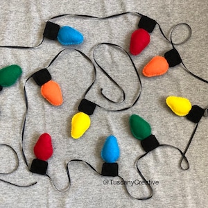 Puede incluir: Una guirnalda de luces navideñas de fieltro en varios colores, incluyendo rojo, verde, azul, amarillo y naranja. Las luces están enhebradas con cinta negra y tienen bases de fieltro negro.