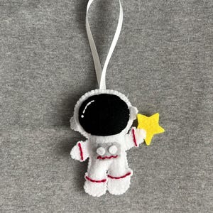 May include: A white felt astronaut ornament with a black felt helmet and red stitching. The astronaut is holding a yellow felt star.