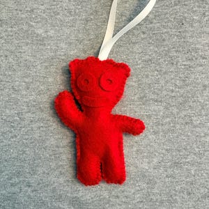 Könnte beinhalten: Handgefertigter roter Filzschmuck in Form einer Cartoonfigur mit weißem Band zum Aufhängen. Der Schmuck hat große kreisförmige Augen und ausgestreckte Arme, vor grauem Hintergrund.
