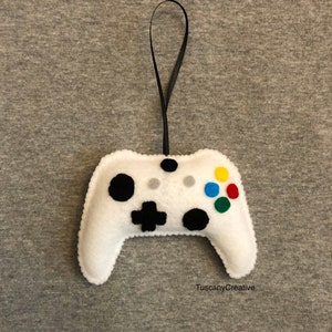 May include: White felt video game controller ornament with black buttons and a black ribbon for hanging. The controller has coloured buttons, including green, blue, red and yellow.