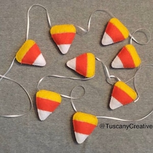 May include: A garland of felt candy corn decorations in yellow, orange, and white. The garland is strung on white ribbon and is perfect for Halloween decor.