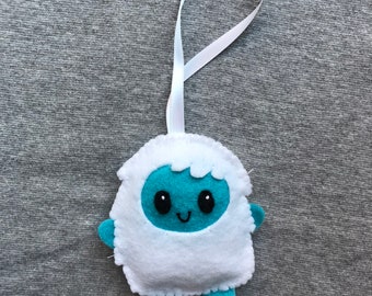 Puede incluir: Un adorno de muñeco de nieve de fieltro blanco con detalles azules y una cara sonriente. El adorno tiene una cinta blanca para colgar.