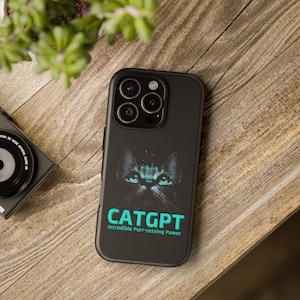 May include: Black phone case featuring a cat graphic with glowing teal eyes. The text "CATGPT" is displayed in teal, with the tagline "Incredible Purr-cessing Power" below. The case is on a wooden surface.