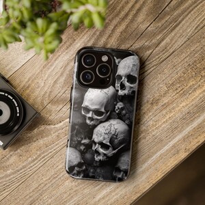 May include: Black and white phone case featuring a detailed skull design. The case displays a pile of skulls in varying sizes, creating a gothic and macabre aesthetic. The case is shown on a wooden surface.