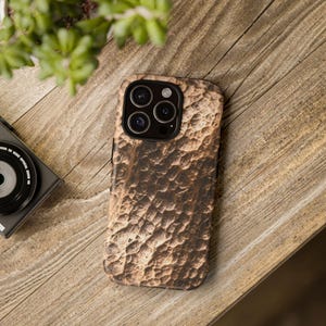 May include: A brown textured phone case with a rugged, bark-like appearance. The case is on a wooden surface, with a camera and greenery in the background. The phone case has a multi-lens camera system.