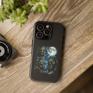 May include: Black phone case featuring a graphic of swamp creatures with a full moon and lightning. The case is on a wooden surface, with a camera and plant in the background. The phone case has a glossy finish.