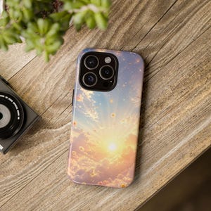 Pode incluir: Uma capa de telemóvel com um design de pôr do sol, com nuvens, um sol brilhante e pequenas flores. A capa está numa superfície de madeira, com uma câmara e uma planta visíveis no fundo.