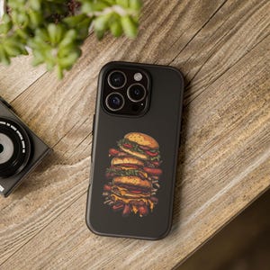 May include: Black phone case featuring a stacked burger illustration with sesame seed buns, lettuce, tomato, and cheese. The design includes visible sausage links. The case is on a wooden surface.