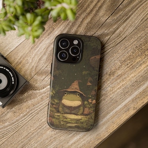 Op de afbeelding: Donkergroene telefoonhoes met een grillige illustratie van een kikker met een tovenaars hoed en vest. De hoes heeft een matte afwerking en een beschermende bumper. De achtergrond bevat paddenstoelen en gebladerte.