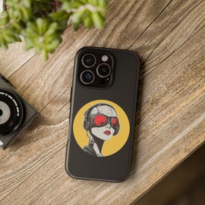 May include: Black phone case featuring a retro-futuristic illustration of a woman with red goggles and headphones on a yellow circle. The phone case is on a wooden surface, with a camera visible in the background.