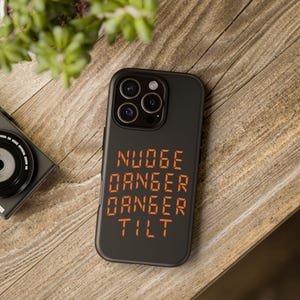 May include: Black phone case with orange text that reads "NUDGE, DANGER, DANGER, TILT." The case is on a wooden surface, with a camera and plant in the background.