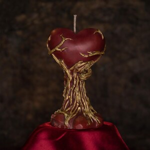 Puede incluir: Una vela en forma de corazón burdeos con una base dorada en forma de árbol. La vela se asienta sobre una superficie de satén rojo sobre un fondo marrón oscuro. La vela tiene una mecha en la parte superior.